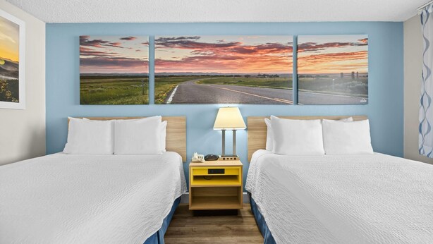 A hotel room with two beds, a nightstand with a lamp, and a wall with three landscape paintings. - Days Inn by Wyndham Lost Hills (Lost Hills)