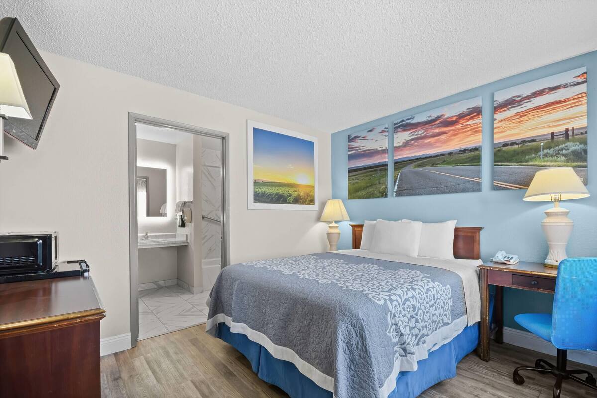 A hotel room with a bed, desk, chair, and artwork on the wall.
