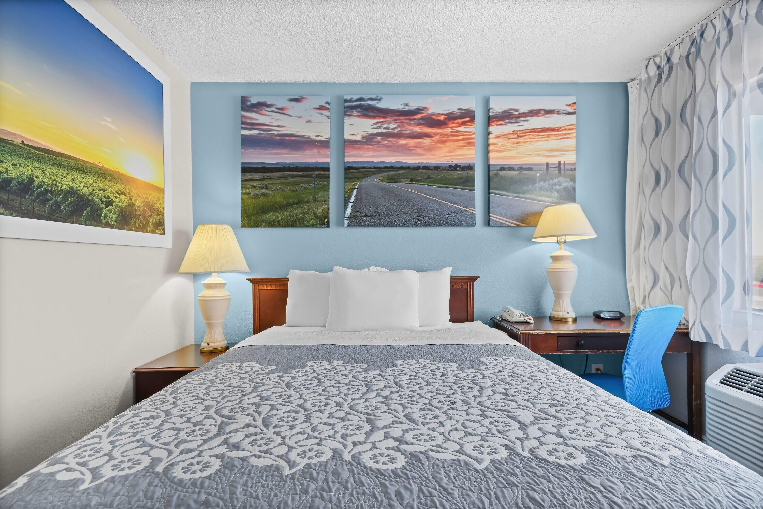 Ein Hotelzimmer mit einem Bett, zwei Nachttischlampen, einem Schreibtisch mit Stuhl und drei großen Landschaftsbildern an der Wand.