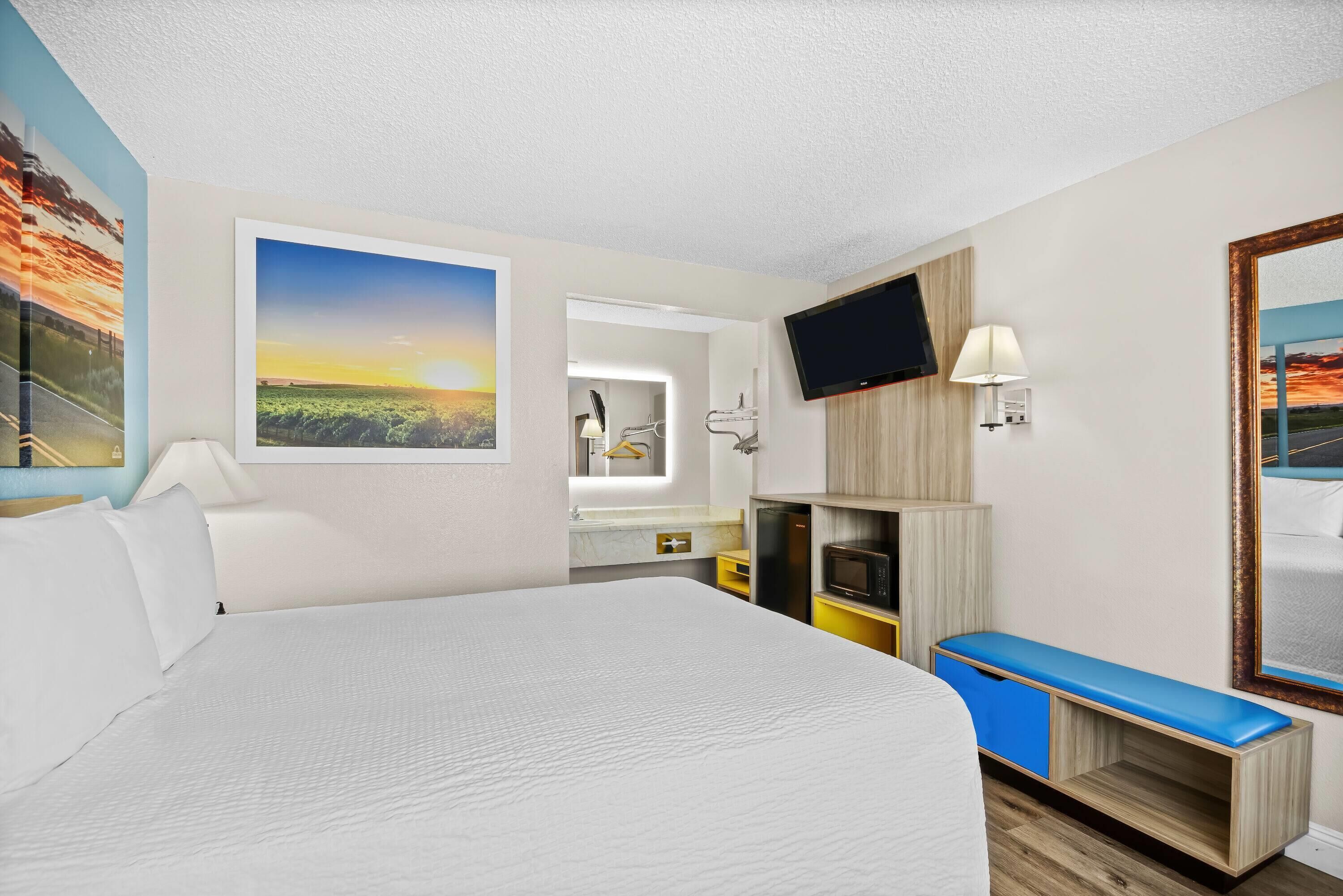 A hotel room with a large bed, a TV mounted on the wall, a desk with a lamp, and a bench.
