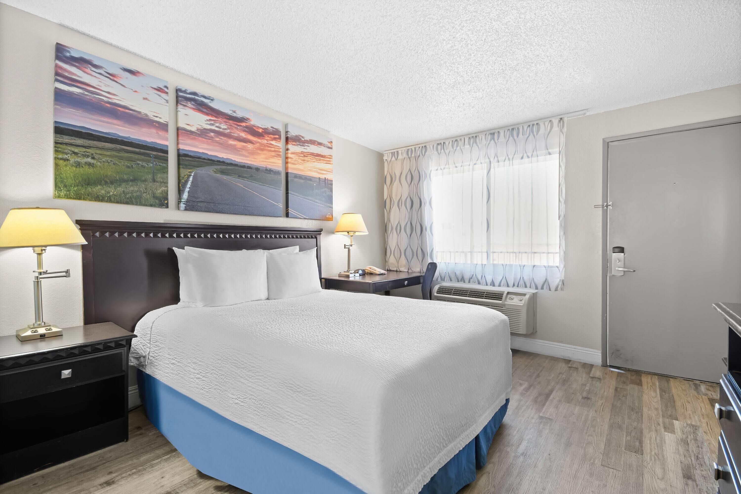 A hotel room with a large bed, two bedside tables with lamps, a window with curtains, and a door.