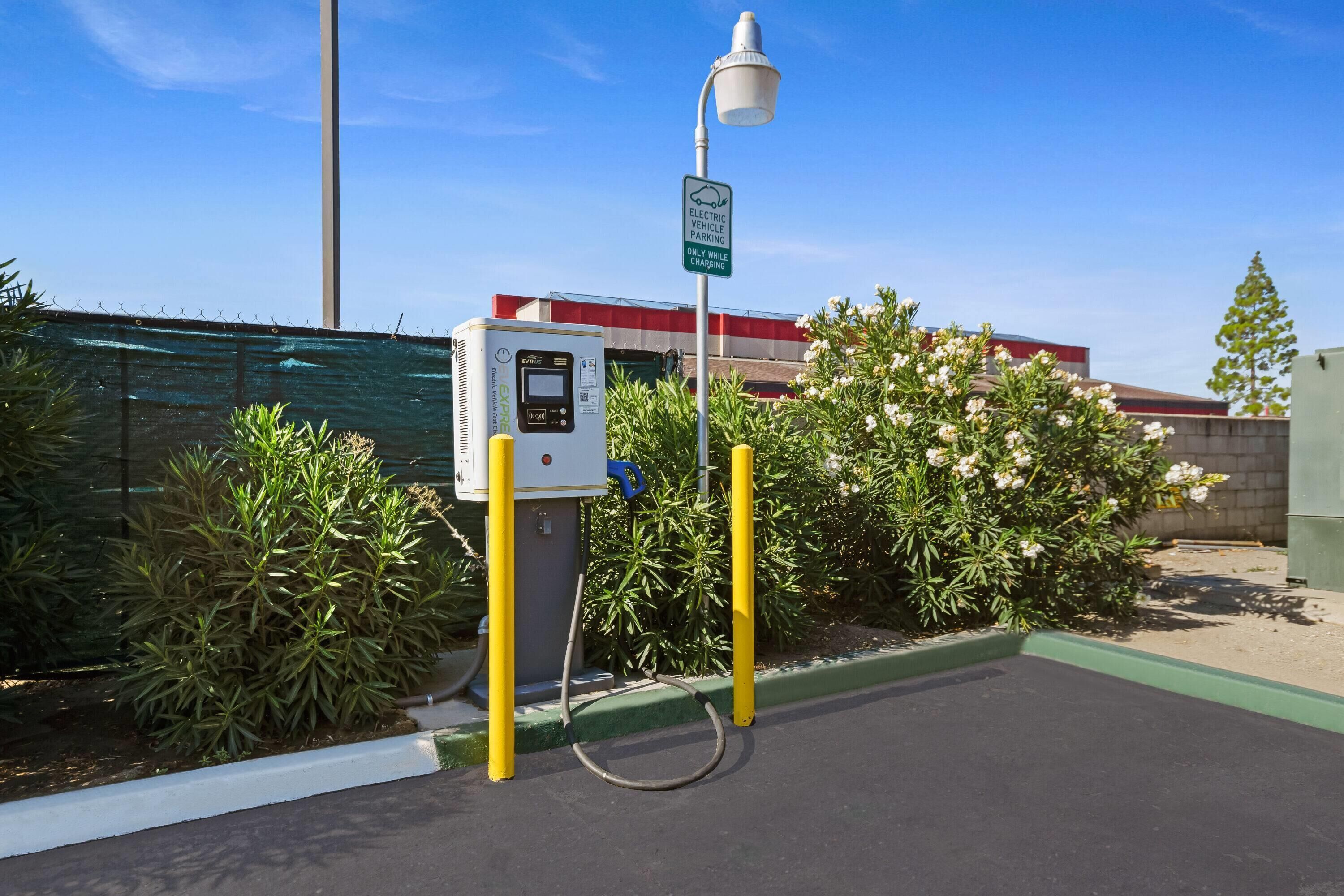 An electric vehicle charging station with a control panel, a sign indicating 'ELECTRIC VEHICLE PARKING ONLY WHILE CHARGING,' and a lamp post.