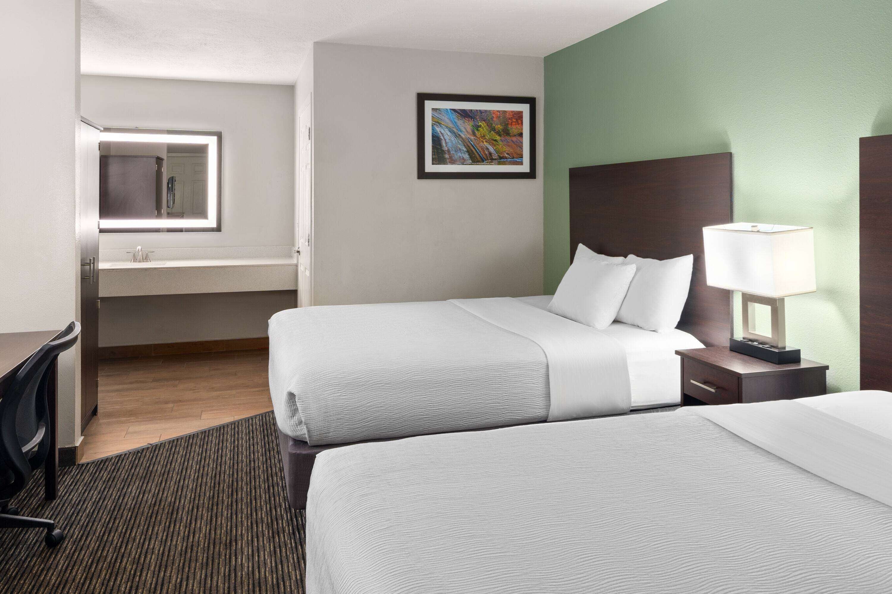 A hotel room with two beds, a desk, a chair, a nightstand with a lamp, a framed picture on the wall, and a window with a mirror.