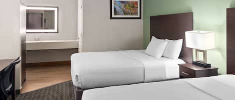 A hotel room with two beds, a desk, a chair, a nightstand with a lamp, a framed picture on the wall, and a window with a mirror.