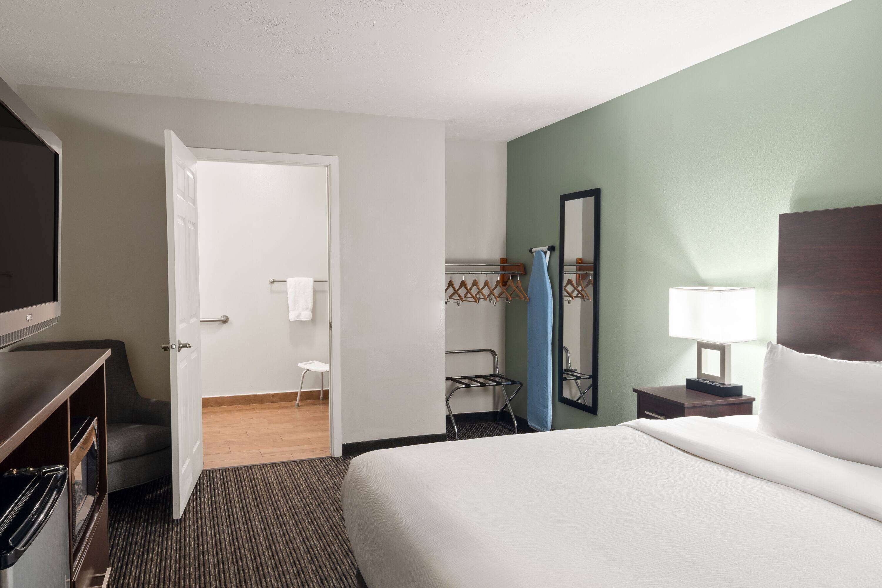 A hotel room with a bed, a TV, a desk with a lamp, a chair, and a bathroom visible through an open door.