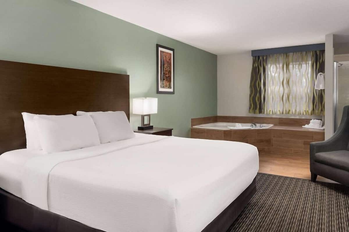 Una habitación de hotel con una cama grande, una mesita de noche con lámpara, un baño con lavamanos y espejo, y una silla.