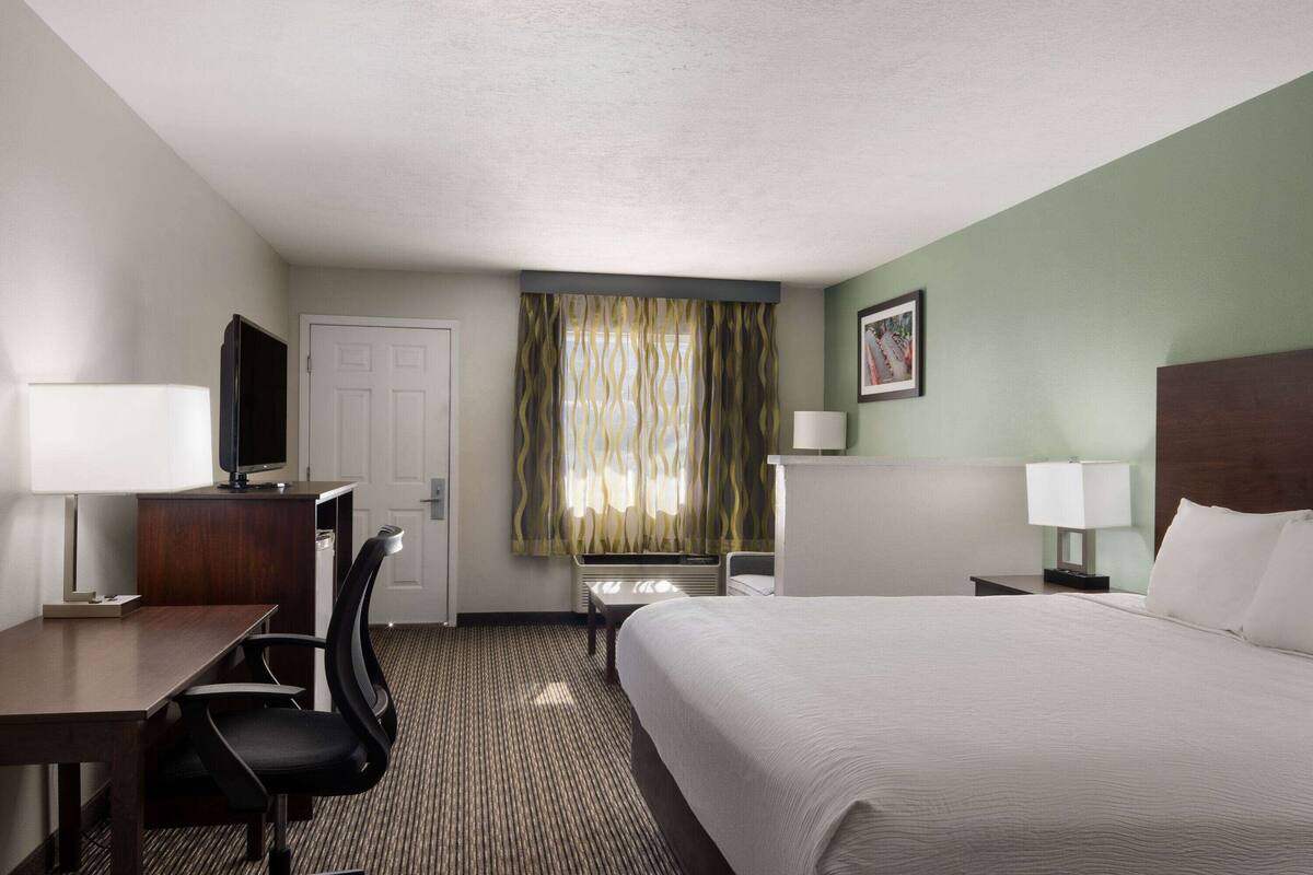 Una habitación de hotel con cama, escritorio, silla, televisor y ventana con cortinas.