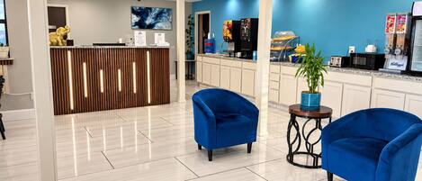 Eine moderne Lobby mit einer Rezeption, zwei blauen Sesseln, einer Kaffeemaschine und einer Pflanze.