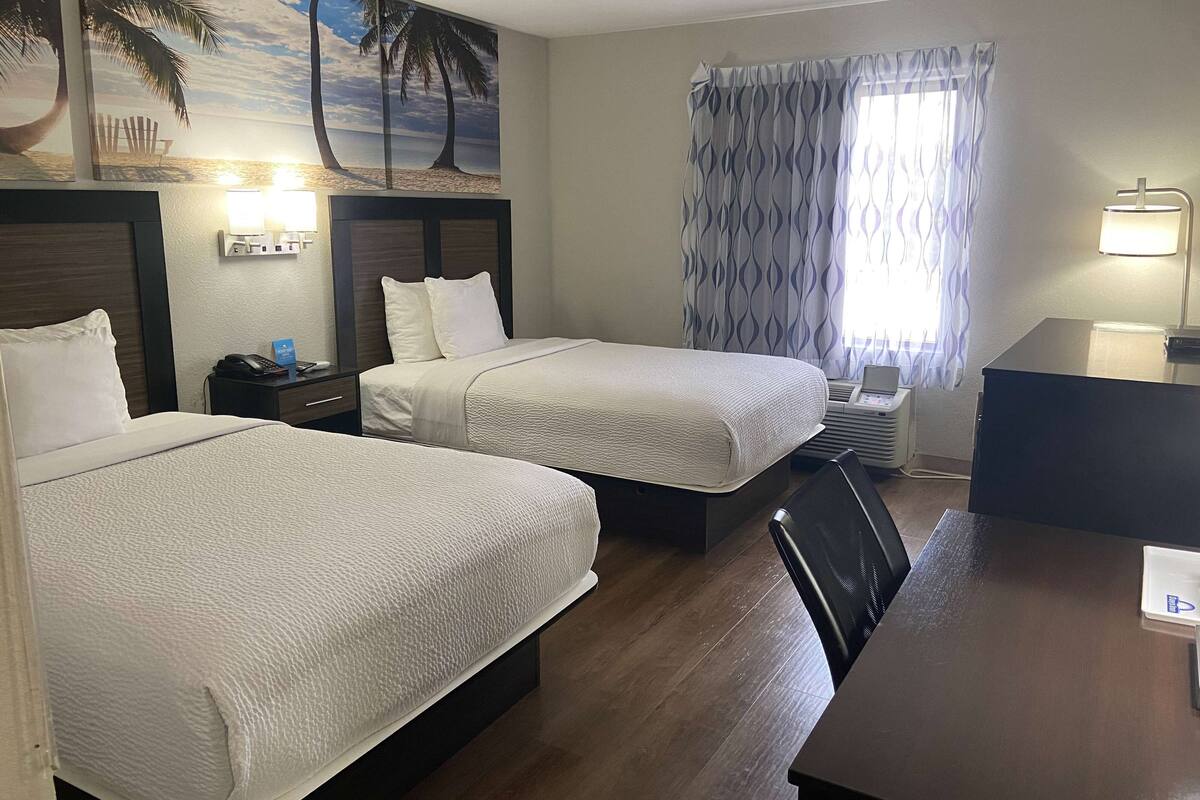 Ein Hotelzimmer mit zwei Betten, einem Schreibtisch, einem Stuhl, einer Lampe und einem Wandbild mit Strandmotiv.