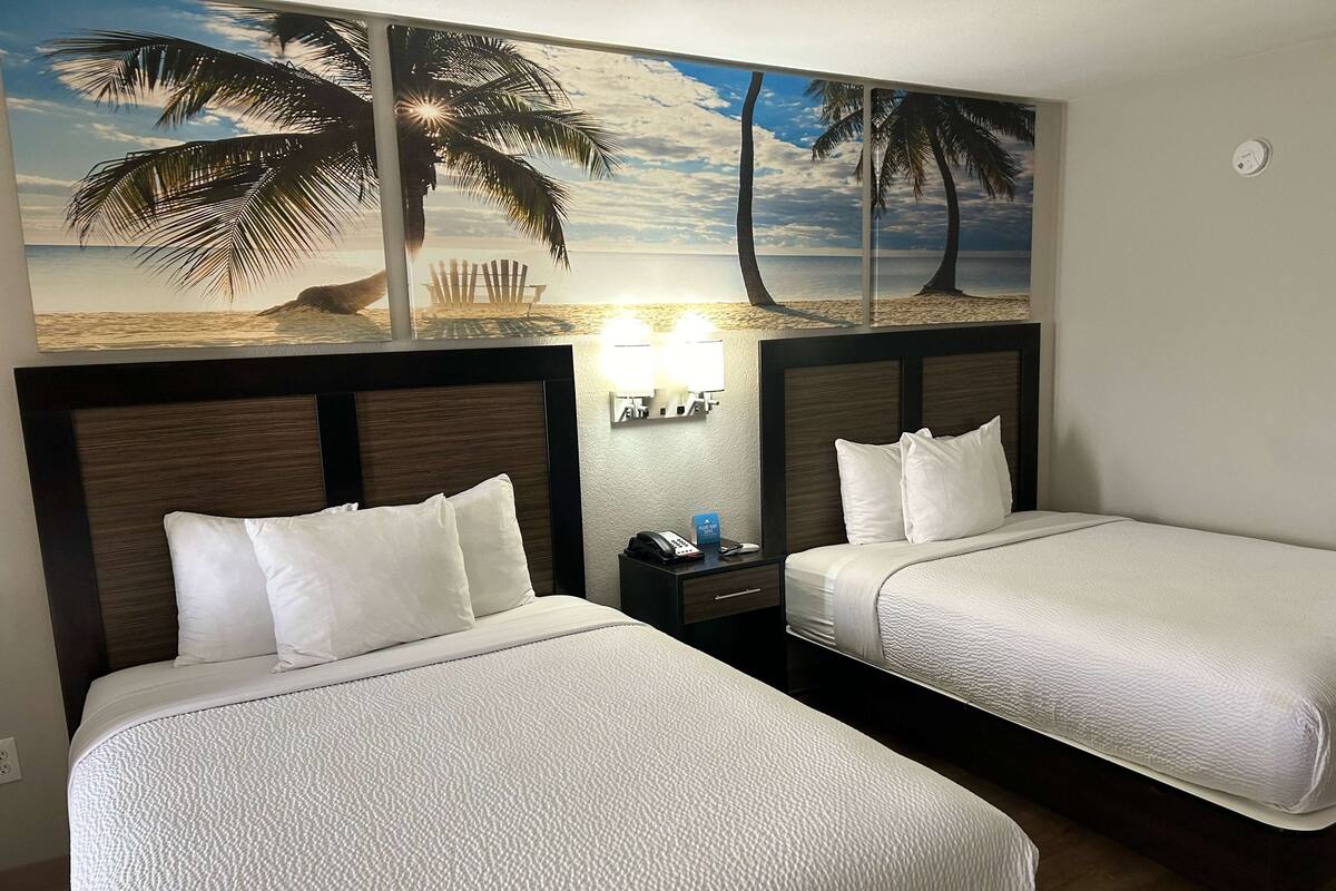 Ein Hotelzimmer mit zwei Betten, einem Nachttisch mit Lampe und einem Wandgemälde mit Strandmotiv.