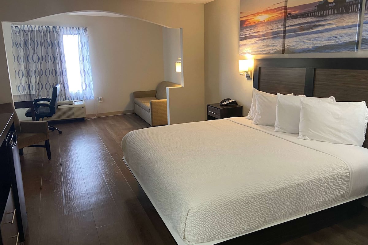 Ein Hotelzimmer mit einem großen Bett, einem Schreibtisch mit Stuhl, einem kleinen Tisch, einem Nachttisch mit Lampe und einem Gemälde, das einen Sonnenuntergang über dem Meer zeigt.