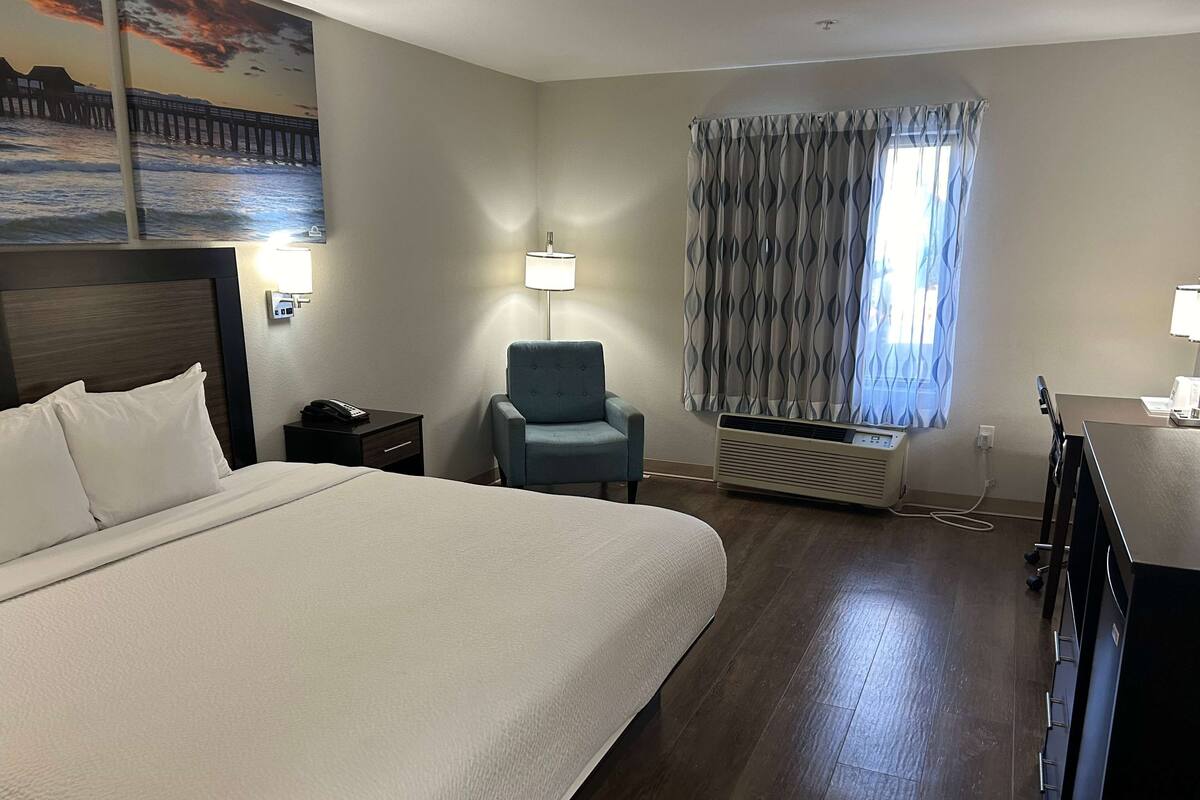 Ein Hotelzimmer mit einem großen Bett, einem Sessel, einem Schreibtisch, einer Lampe, einem Fenster mit Vorhängen und einer Wand-Klimaanlage.