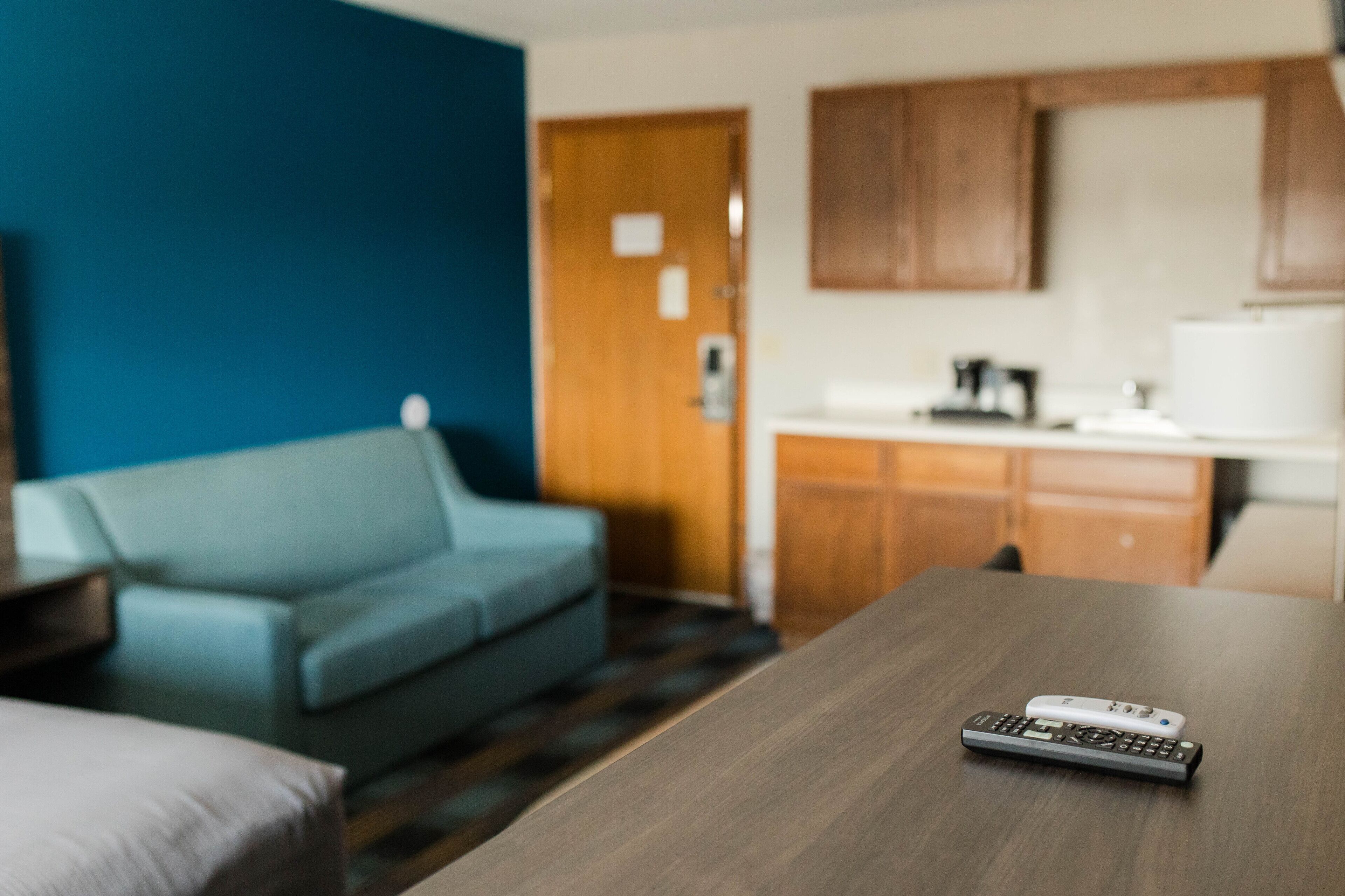 A hotel room with a wooden table, a remote control, a blue sofa, and a kitchen area with wooden cabinets and a refrigerator.