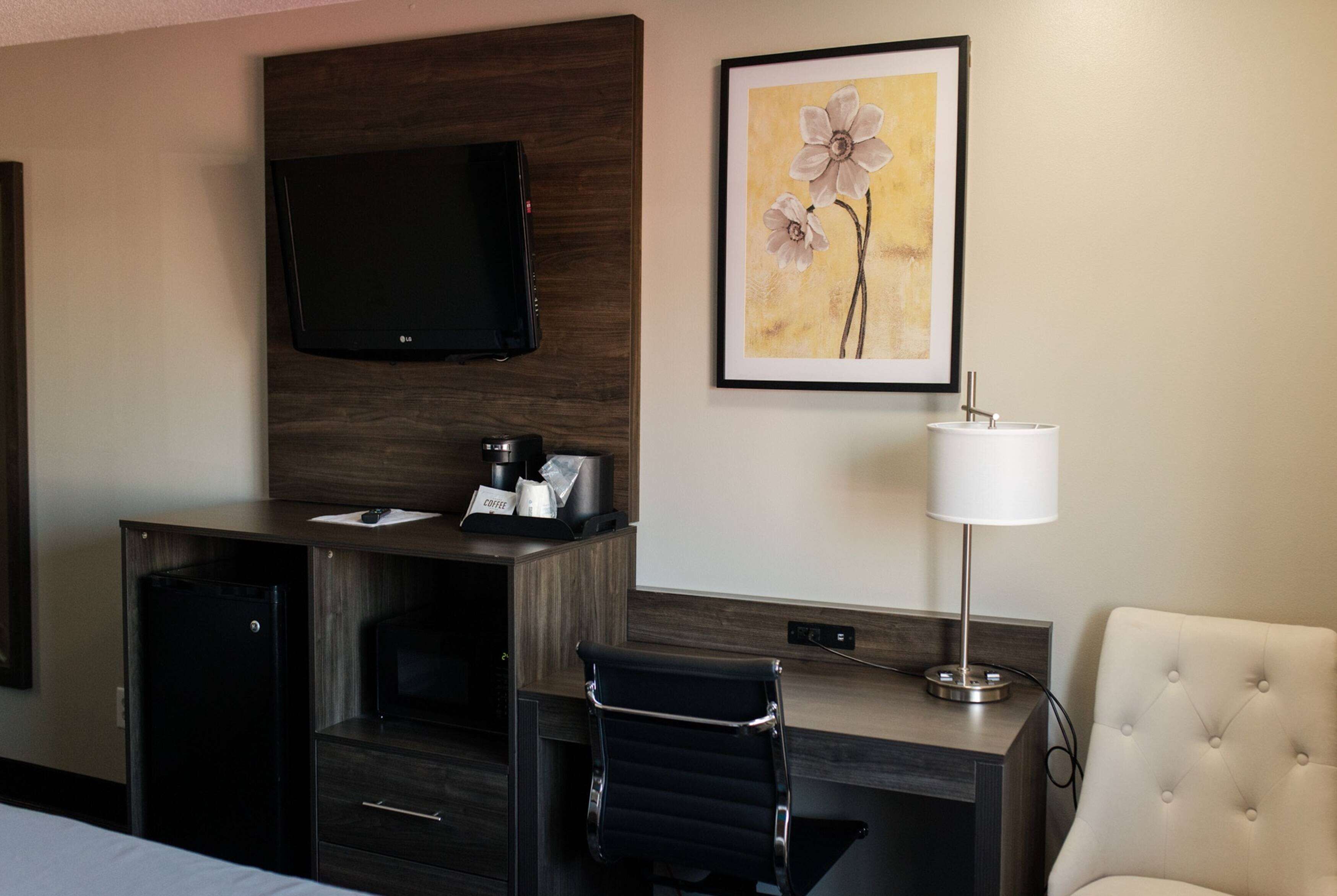 A hotel room with a flat-screen TV mounted on the wall, a coffee maker, a framed floral artwork, a lamp, a desk with a chair, and a bed.