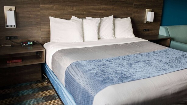 A hotel room with a wooden headboard, a large bed with white bedding, a blue textured throw blanket, a nightstand with a lamp, and a wall-mounted light fixture. - Travelodge by Wyndham Escanaba (Escanaba)