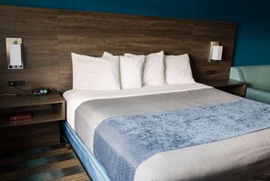 A hotel room with a wooden headboard, a large bed with white bedding, a blue textured throw blanket, a nightstand with a lamp, and a wall-mounted light fixture.