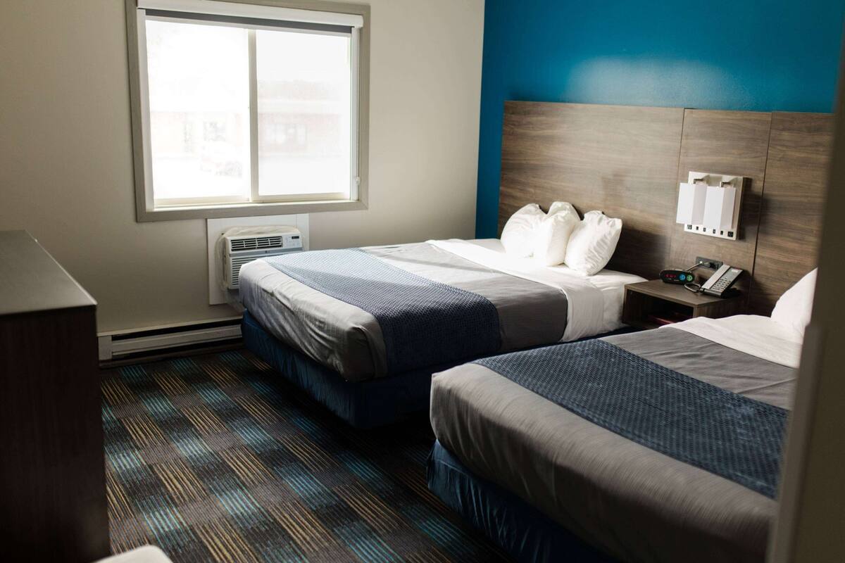 Habitación de hotel con dos camas, una ventana, un equipo de aire acondicionado, una mesita de noche con un teléfono y un interruptor de luz en la pared.