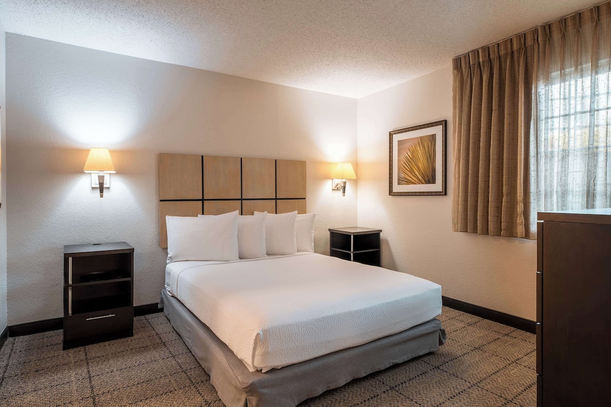 A hotel room with a large bed, two bedside tables, a wall-mounted lamp, a framed picture, and a window with curtains.