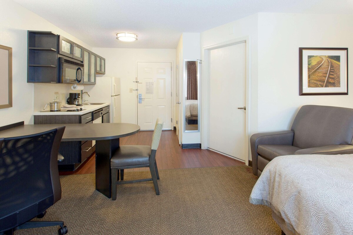 Une chambre d’hôtel de taille réduite comprenant un lit, un bureau avec une chaise, une kitchenette et une porte donnant accès à une salle de bain.