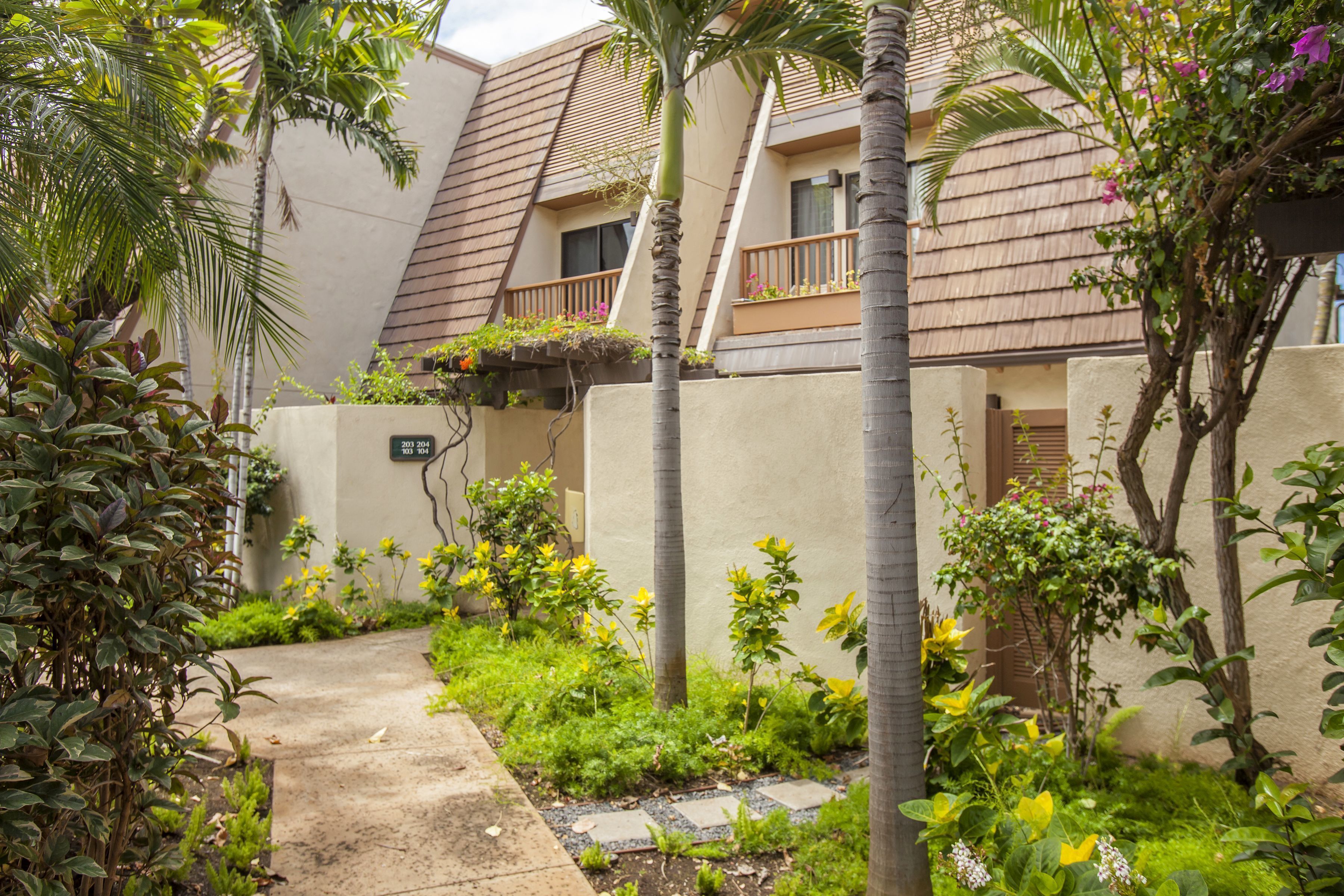 A residential area with a paved pathway, lush greenery, and a building with a sloped roof and balconies.