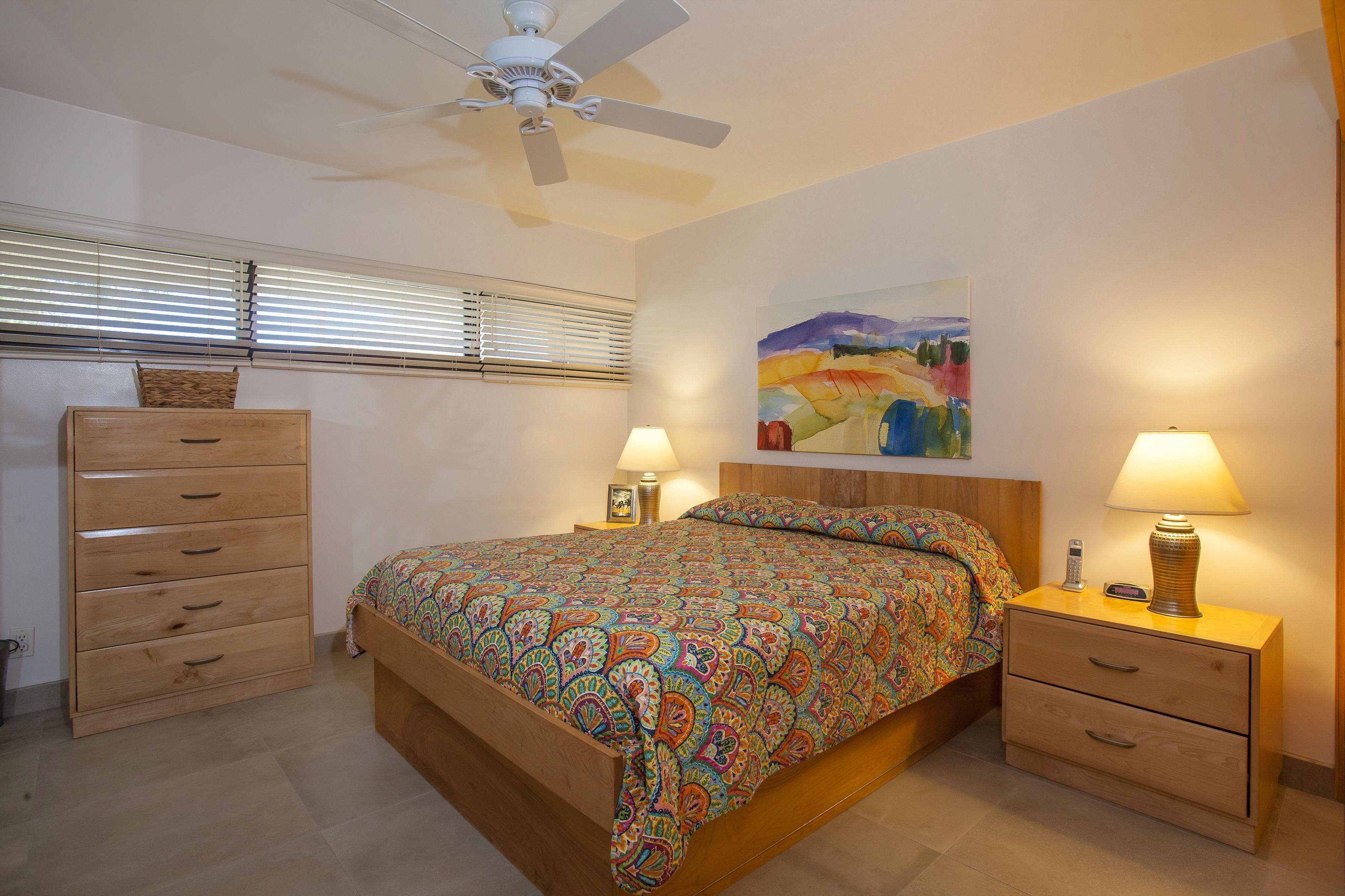 A bedroom with a bed, bedside tables, a ceiling fan, a window with blinds, and a painting on the wall.
