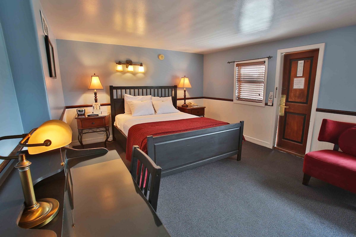 A hotel room with a bed, two bedside lamps, a red chair, a wooden door, and a window with blinds.