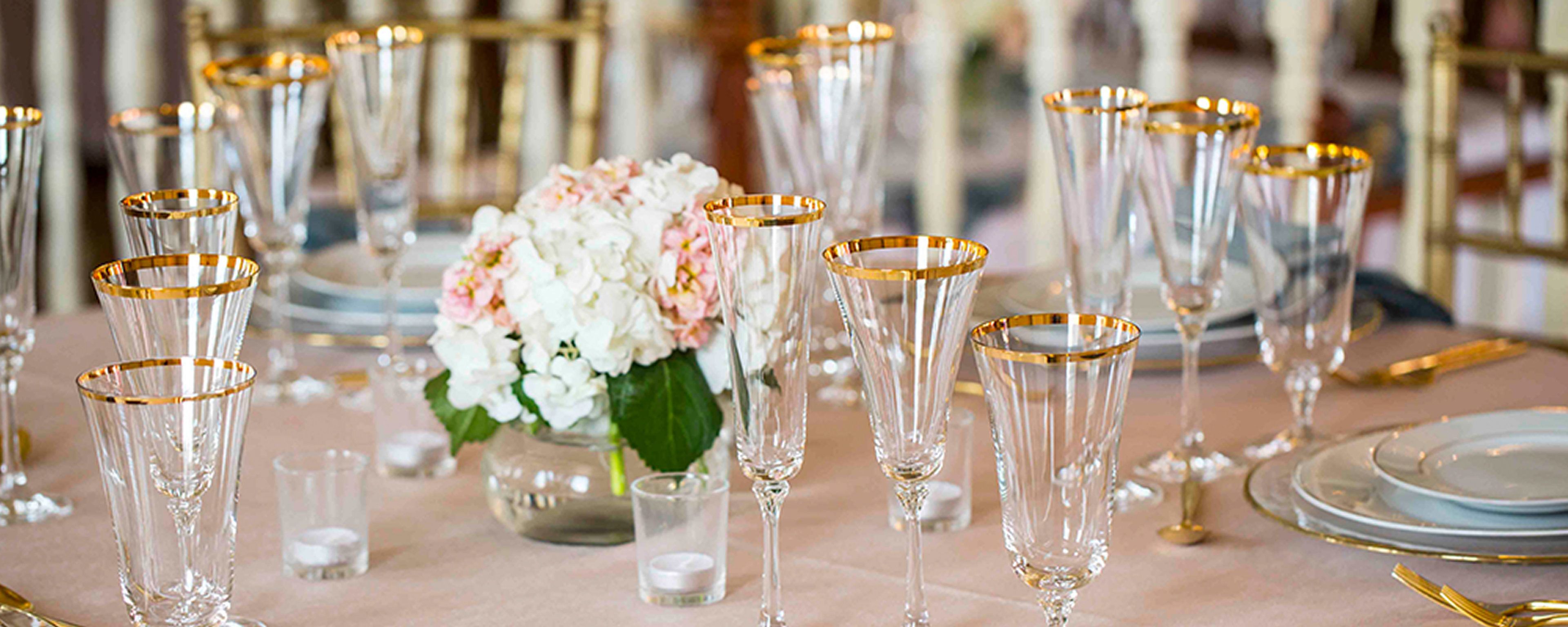Une table dressée avec de beaux verres et des bordures dorées, un vase rempli de fleurs et une nappe rose.