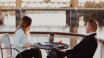 Two people seated at a table with a laptop and drinks, overlooking a river with a bridge in the background.
