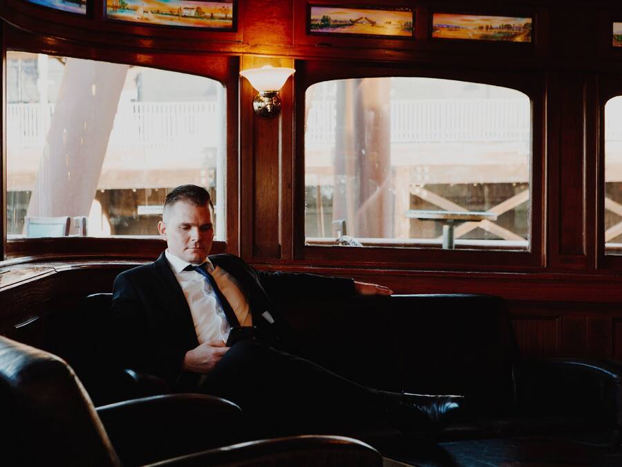 A man seated in a leather armchair inside a room with wooden paneled walls and large windows.