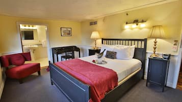 Habitación de hotel con cama, escritorio, silla, mesita de noche y baño visible a través de una puerta abierta.
