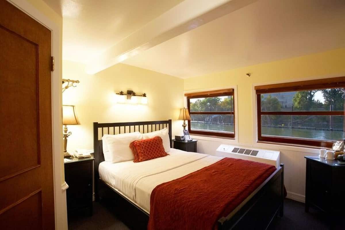A hotel room with a bed, bedside lamps, a window with a view, and a thermostat control panel.