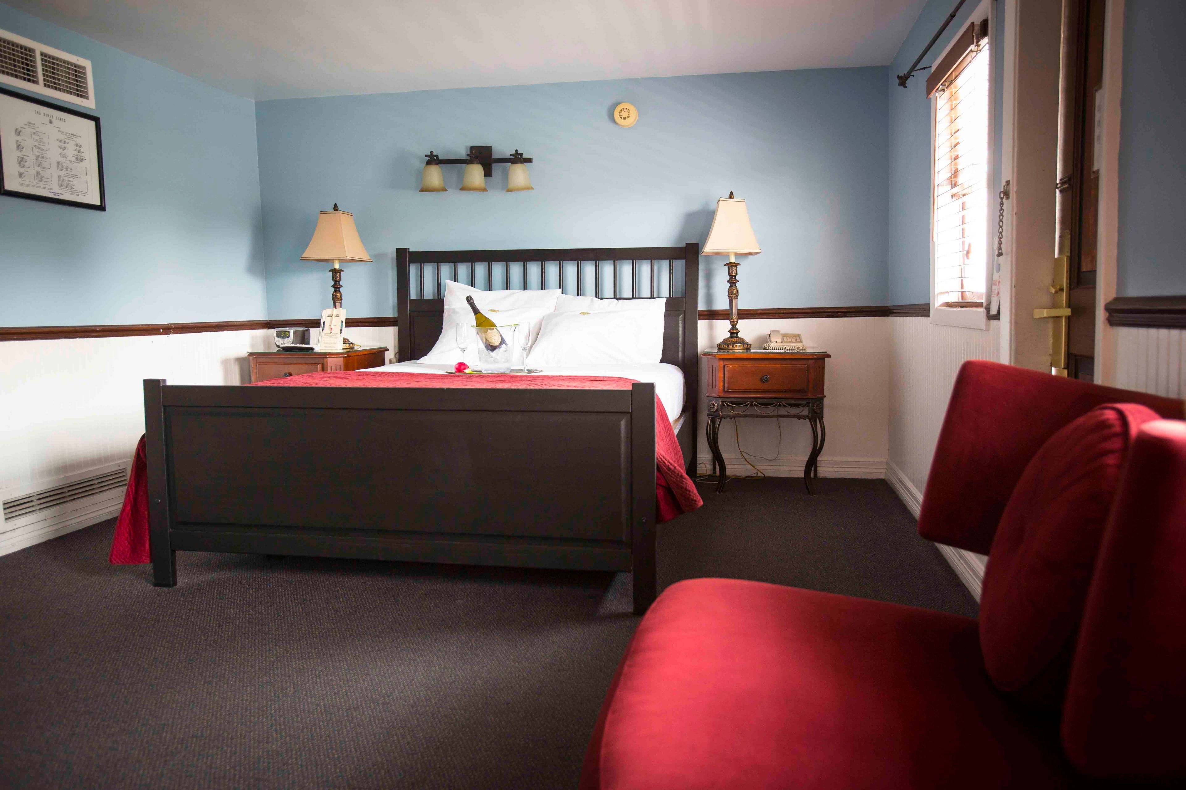 A hotel room with a bed, two bedside tables, a red chair, and a window with blinds.
