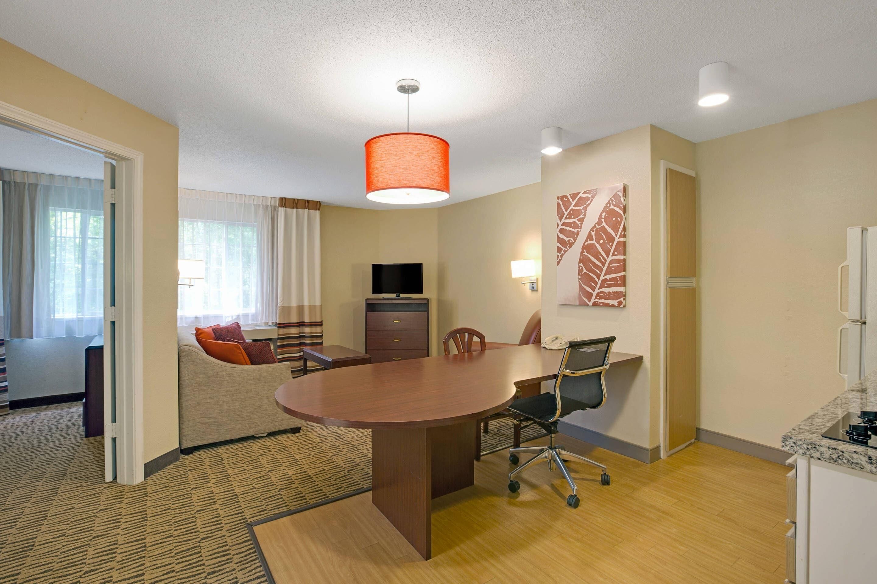 A hotel room with a wooden dining table, a sofa, a TV, and a kitchen area.