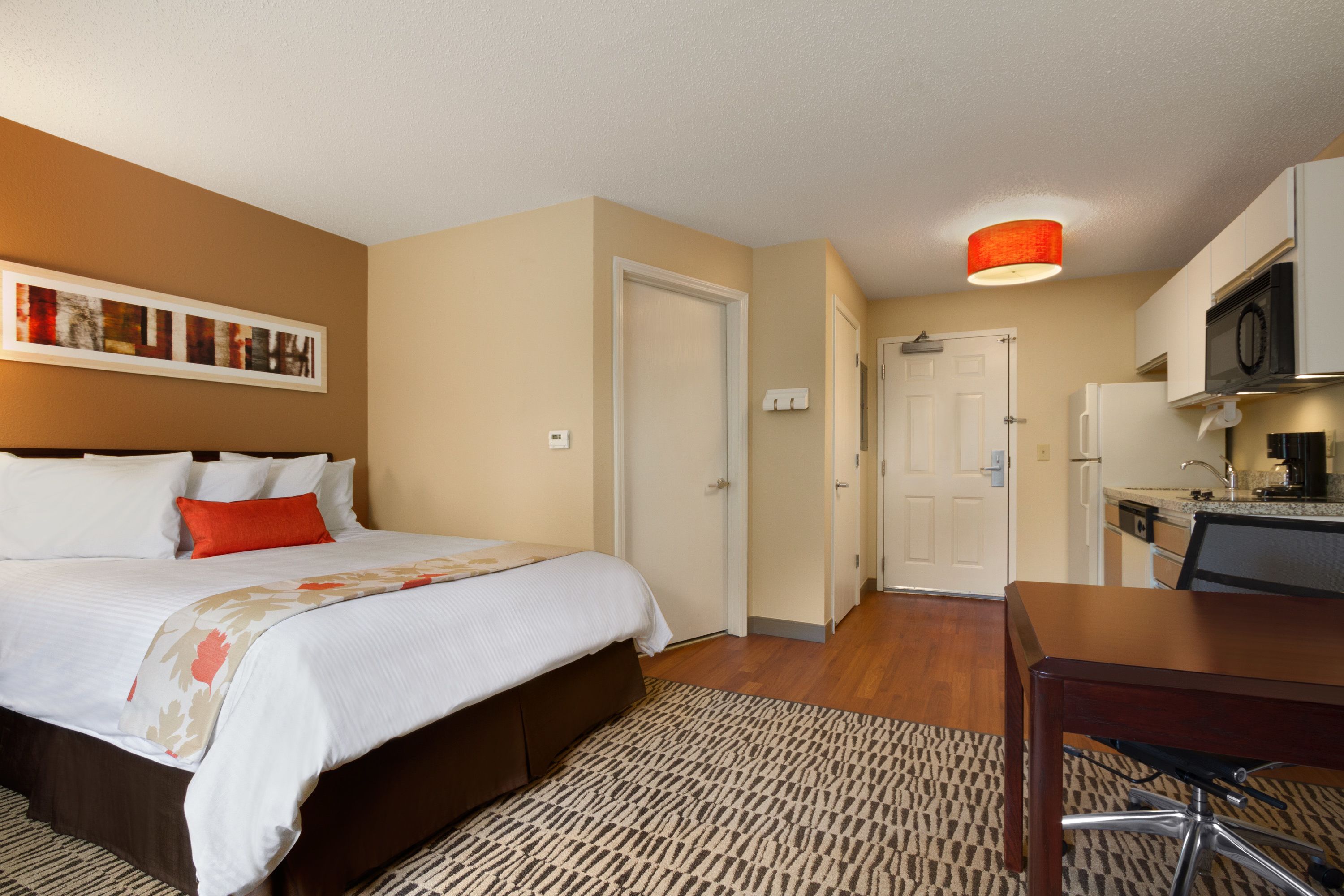 Quarto de hotel com cama, escrivaninha e cozinha compacta.