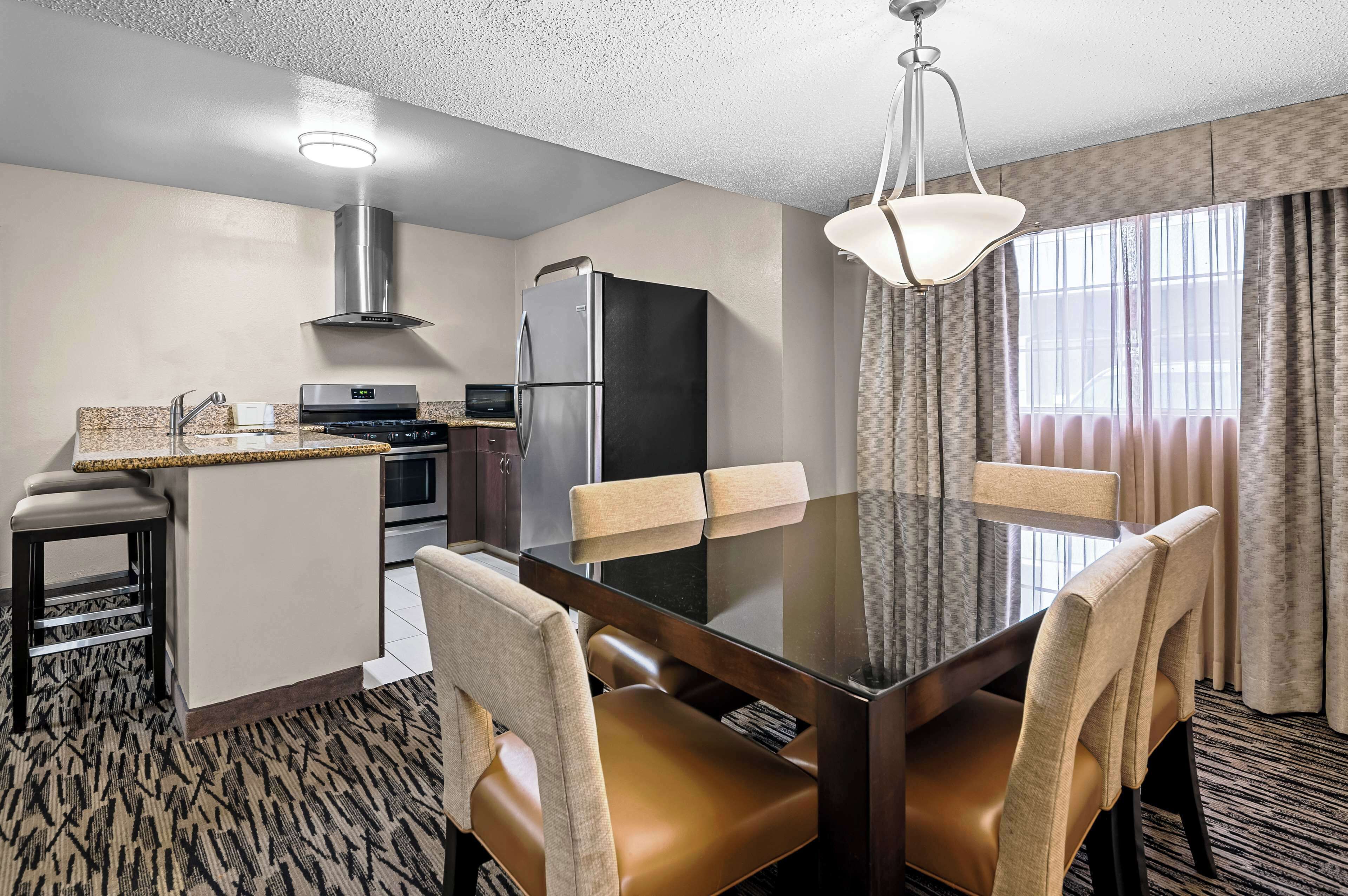A hotel room with a dining table, chairs, a kitchen area with a refrigerator, and a large window with curtains.