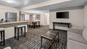 Habitación de hotel con kitchenette, zona de comedor con mesa y sillas, sofá, televisor montado en la pared y alfombra estampada.
