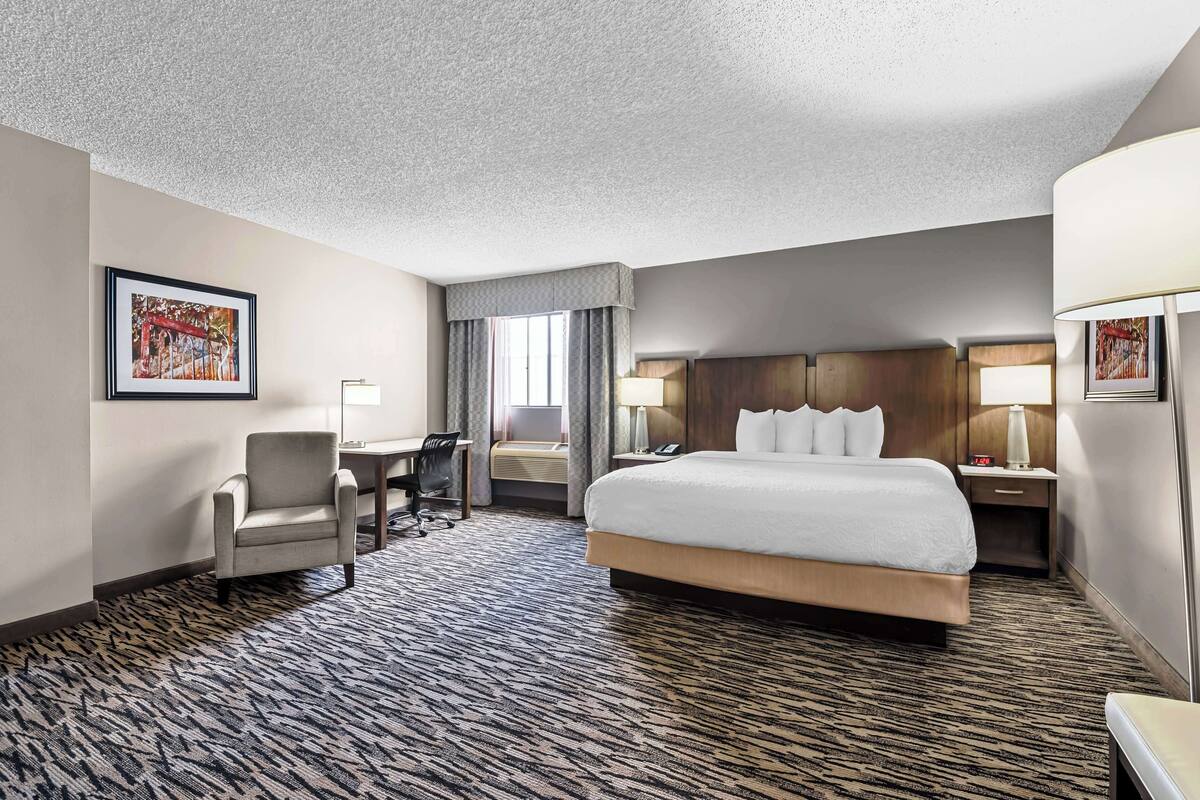 A hotel room with a large bed, a desk with a chair, a small table, and a lamp.