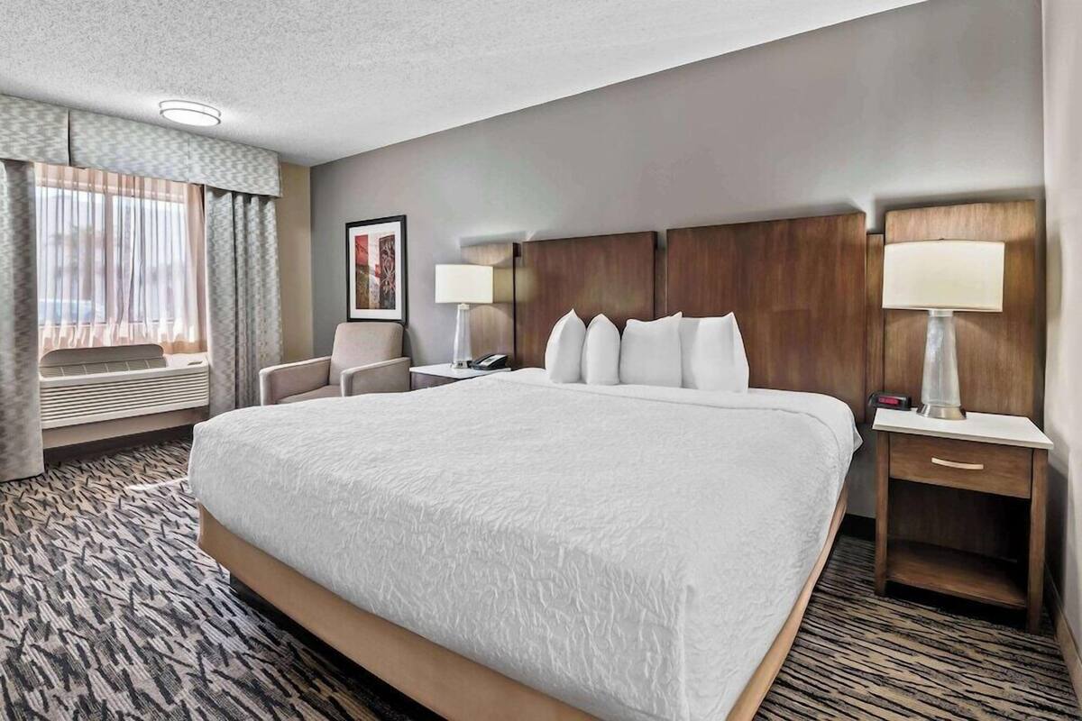 A hotel room with a large bed, two bedside tables with lamps, a chair, a desk, and a window with curtains.