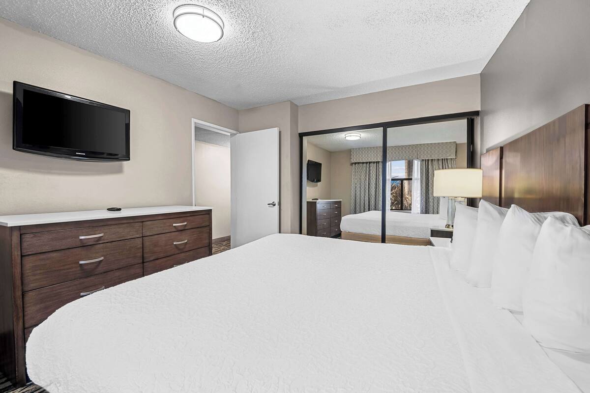 A hotel room with a large bed, a flat-screen TV mounted on the wall, a wooden dresser, and a sliding glass door leading to a balcony.