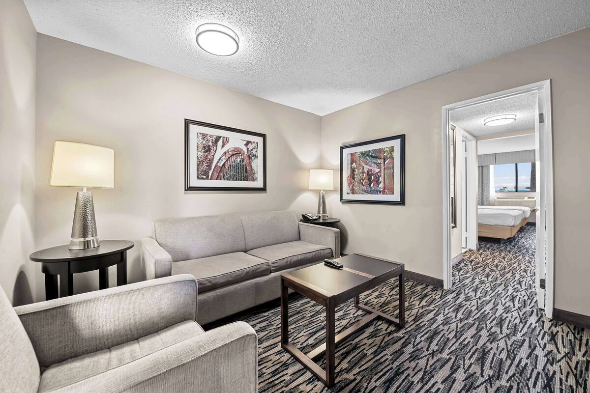 A hotel room with a grey sofa, a small table, two lamps, and framed pictures on the walls.