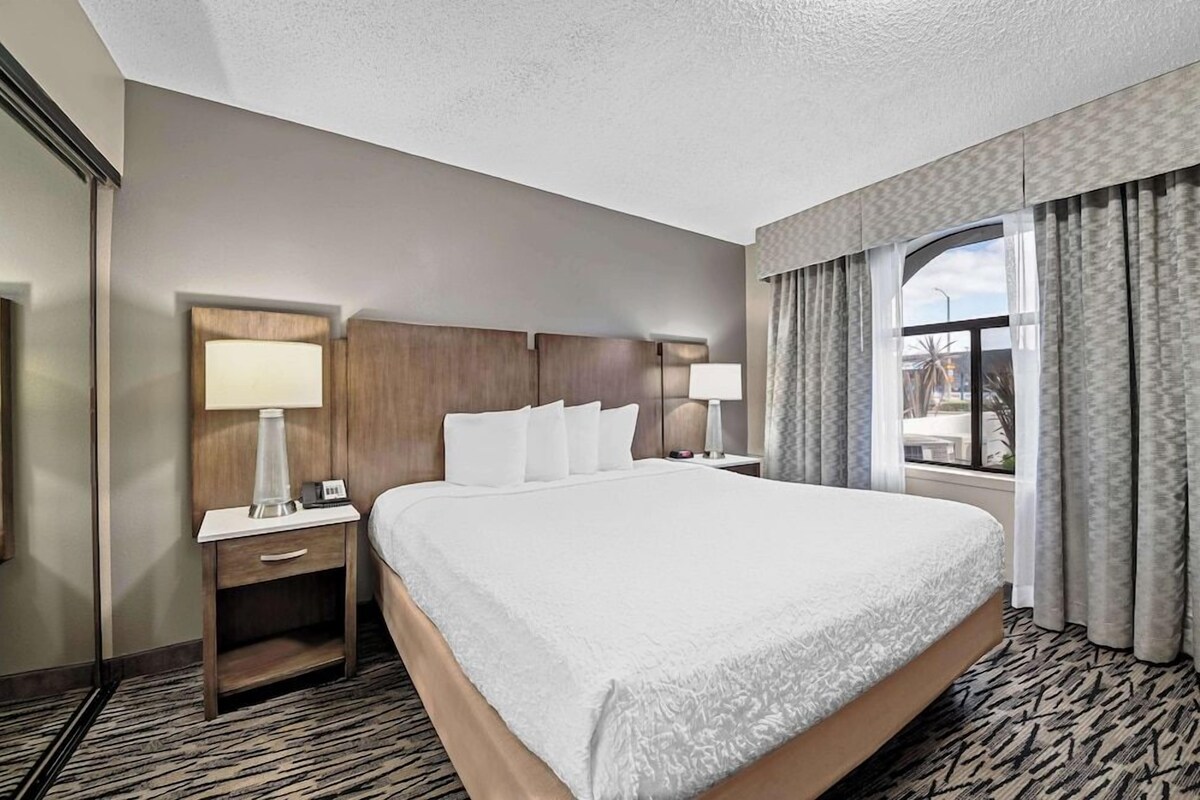 A hotel room with a large bed, two bedside tables with lamps, a nightstand, a TV, and a window with curtains.