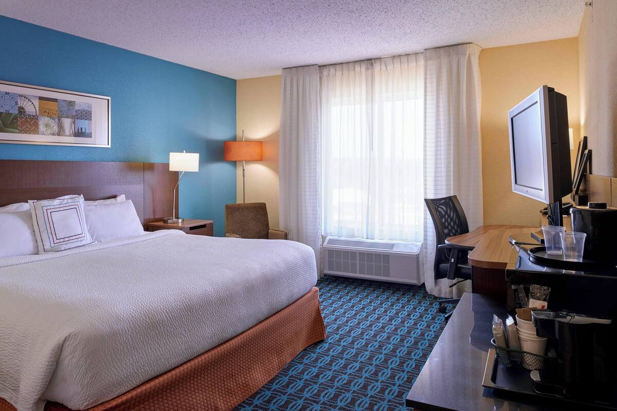 Habitación de hotel con una cama grande, un escritorio con computadora, una silla, televisor y ventana con cortinas.