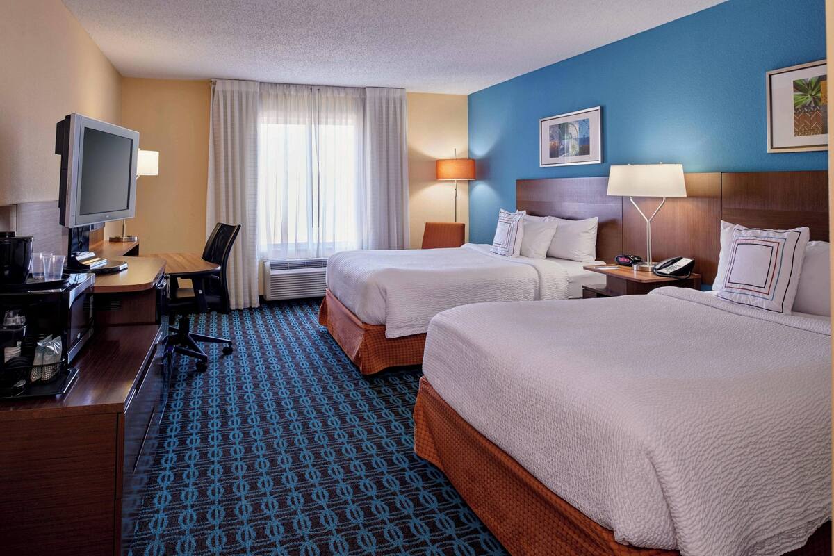 Una habitación de hotel con dos camas, un escritorio con computadora, un televisor y una ventana con cortinas.