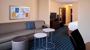 A hotel room with a grey sofa, two small round tables, a TV, and a bed. - Fairfield by Marriott Kalamazoo West (Kalamazoo)