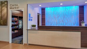 A reception area with a wooden reception desk, a 'Corner Market' sign, and a wall with a tree branch pattern. - Fairfield by Marriott Kalamazoo West (Kalamazoo)