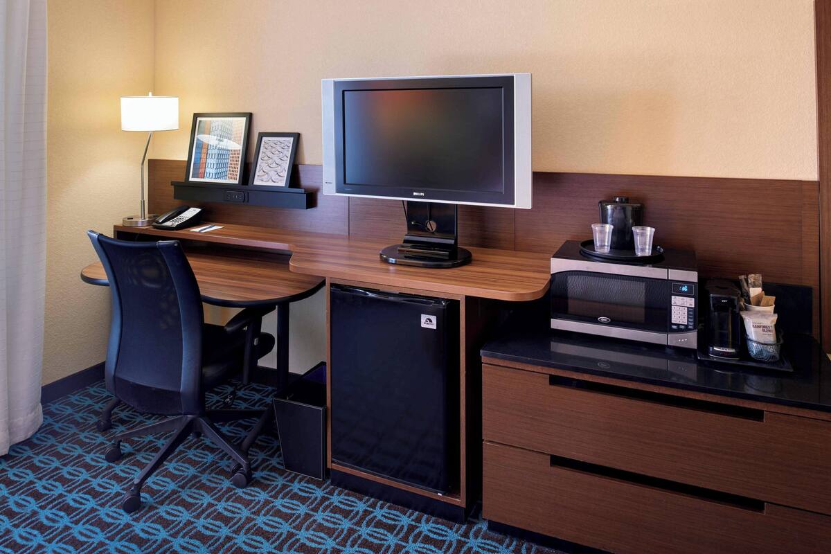 Habitación de hotel pequeña con escritorio, monitor de computadora, refrigerador, microondas, cafetera y una silla.