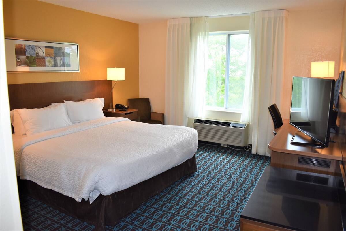 A hotel room with a large bed, a desk with a computer, a TV, a window with curtains, and a painting on the wall.