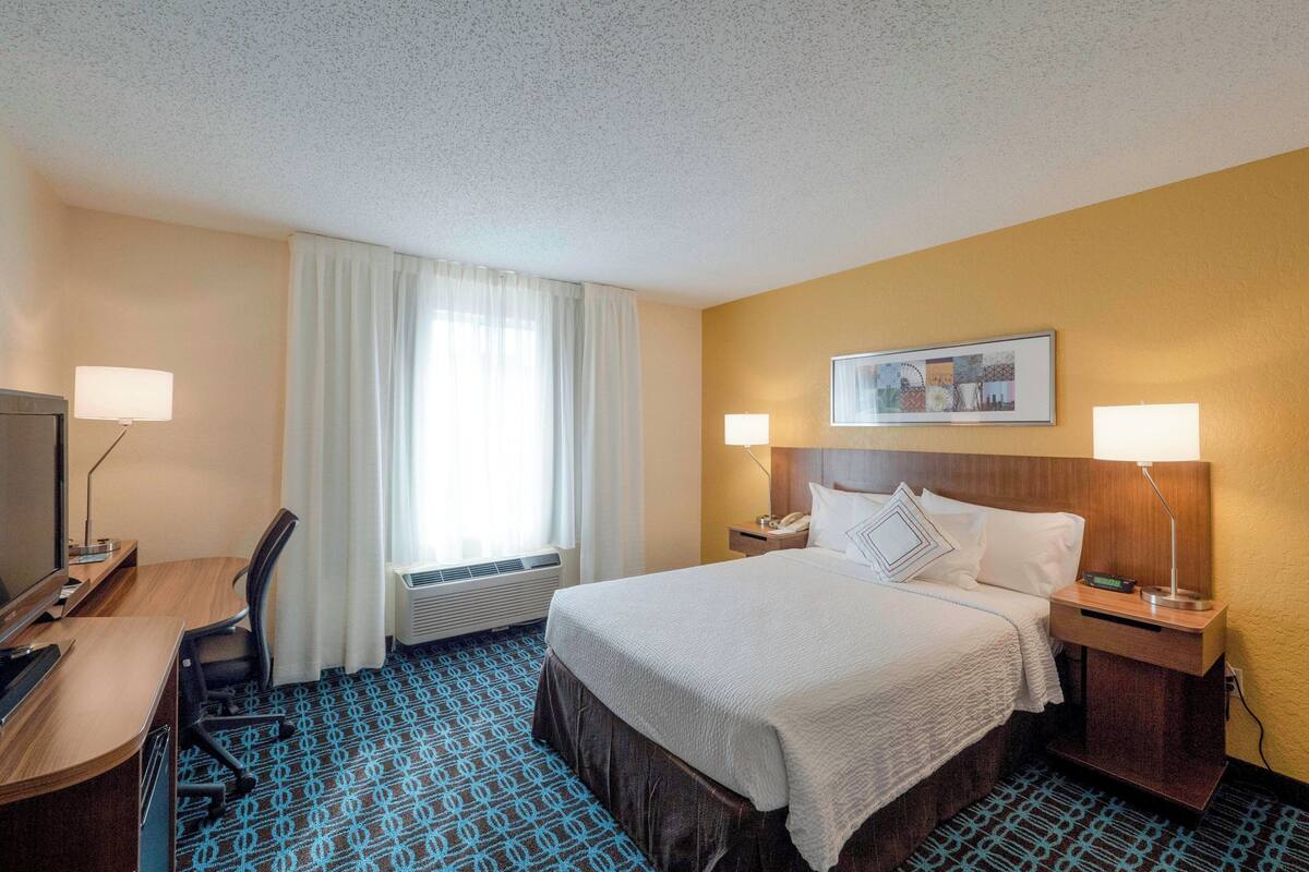 A hotel room with a bed, two bedside tables, a desk, a chair, a TV, and a window with curtains.