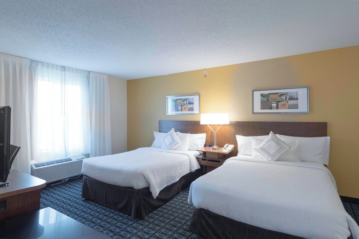 A hotel room with two beds, a desk, a chair, a lamp, and a TV.