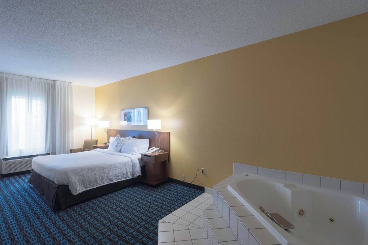 A hotel room with a large bed, a bedside table with a lamp, a wall-mounted TV, and a separate bathroom with a bathtub.